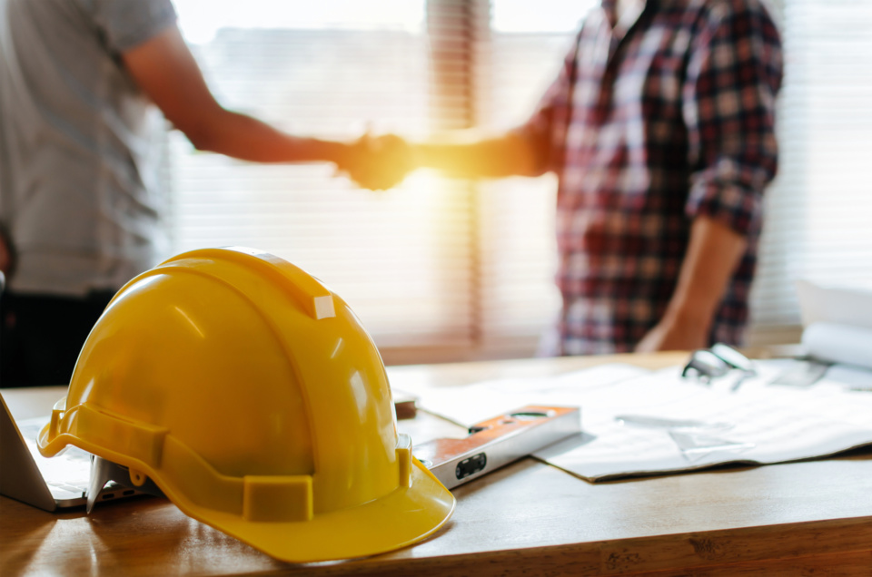 Yellow safety helmet on workplace desk with construction worker team hands shaking greeting start up plan new project contract in office center at construction site partnership and contractor concept 1153235950 2131x1411